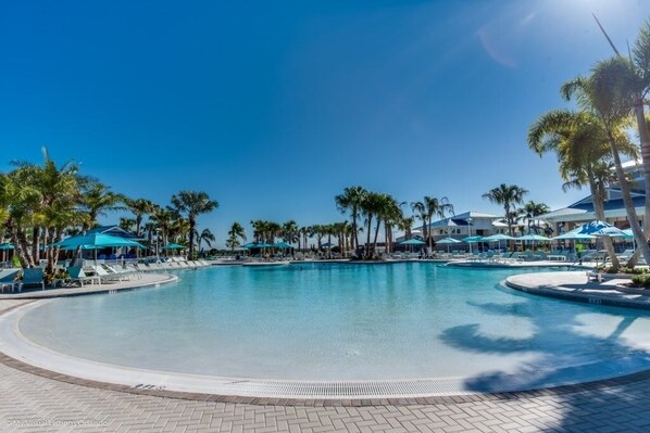 Una gran piscina rodeada de palmeras y sombrillas azules, con sillones de descanso y un cielo azul despejado.