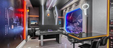 A modern room with a large screen displaying a space-themed image, a sleek black table with chairs, and a unique wall design with geometric patterns and a neon light.