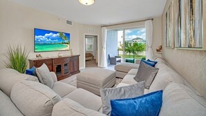 A living room with a large flat-screen TV, a beige sofa, and a view of a balcony with a cityscape.