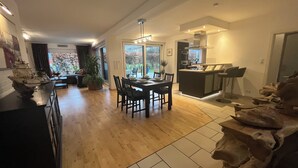 Een ruime woonkamer met houten vloer, een eettafel met stoelen, een keuken met een centraal eiland, en een groot raam met uitzicht op de omgeving.