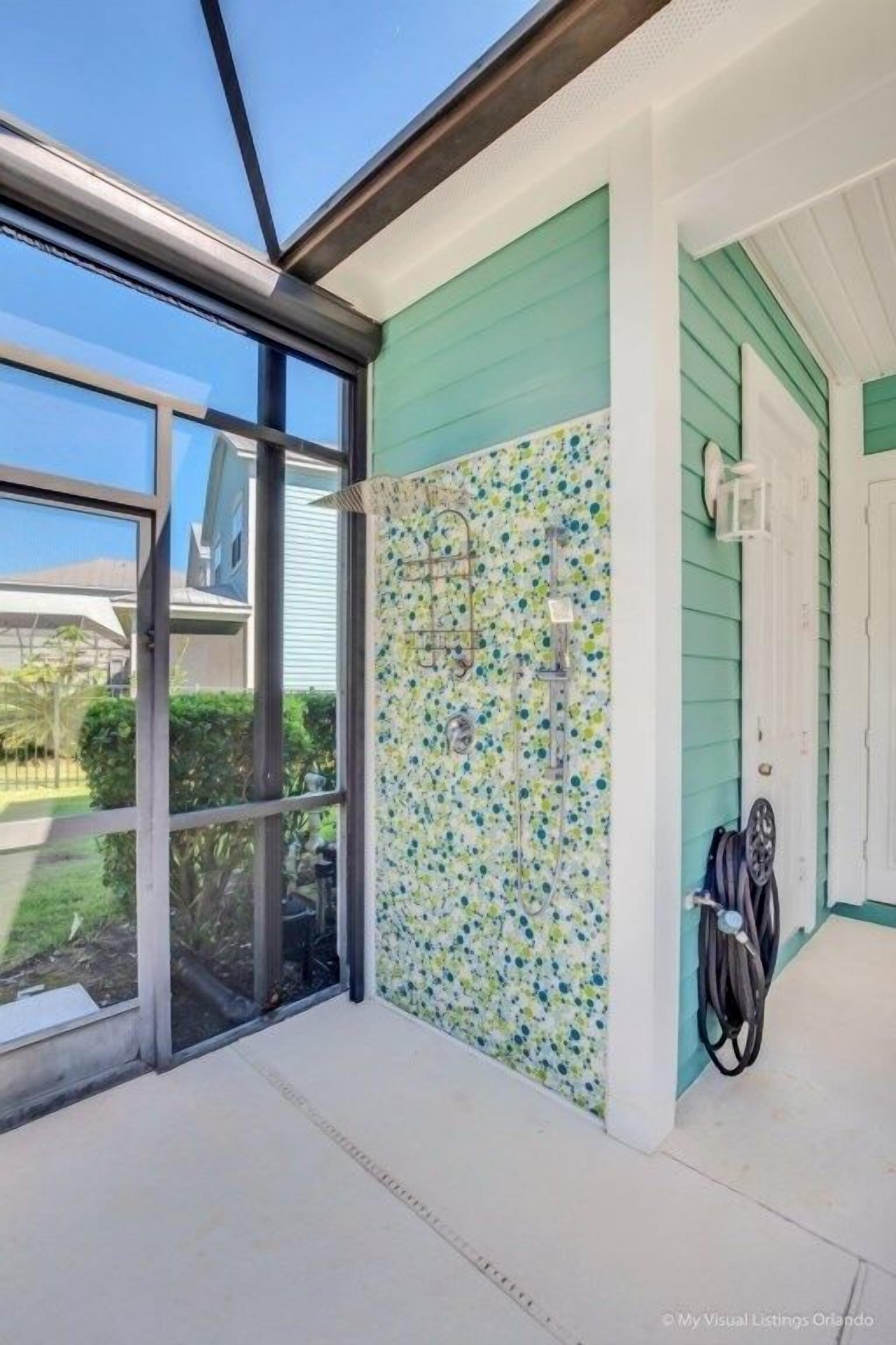 Un espace extérieur abrité, doté d’une porte vitrée, d’une douche avec un mur de carrelage à motifs floraux et d’un tuyau d’arrosage posé sur le sol.