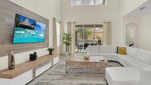 A spacious living room with a large flat-screen TV mounted on the wall, a white sectional sofa, a wooden coffee table, and a large window offering a view of the outdoors.