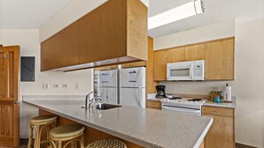 A kitchen with wooden cabinets, a granite countertop, a microwave, a stove, and two wicker stools. - Maui Sunset A412 1 Bedroom Condo (Kihei)