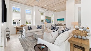 Un salon spacieux, doté de meubles blancs, d’un plafond en bois et de grandes fenêtres.