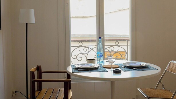 A dining area with a round table, two chairs, a lamp, and a view of the street through a window.