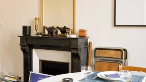 A dining area with a fireplace, a table set with plates, glasses, and a candle. There is a chair with a woven backrest and a book on the table.