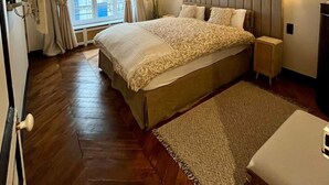 A modern bedroom with a large bed, a headboard with geometric patterns, bedside lamps, and a view of the city through a window with curtains.
