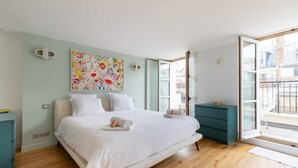 Un dormitori amb un llit gran, una obra d'art colorida a la paret, un aparador verd mar i un balcó amb vista a edificis.