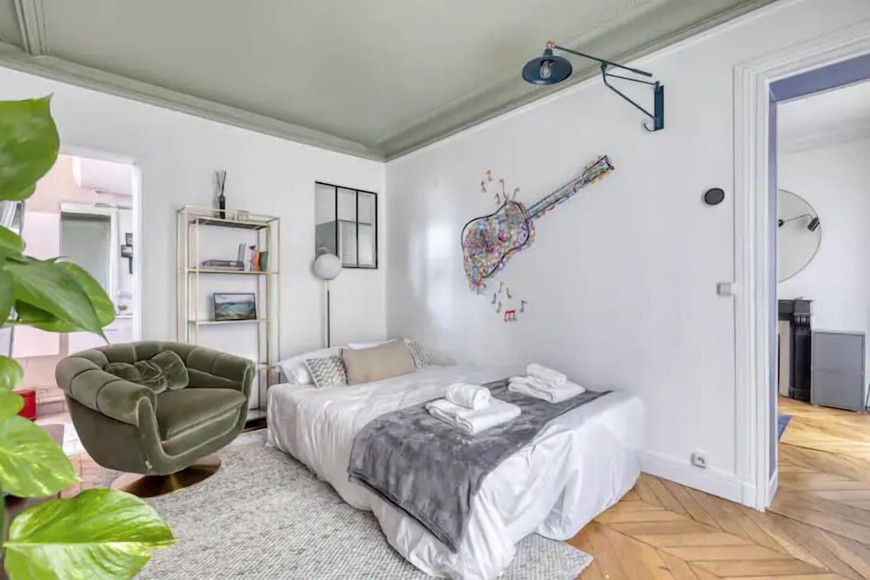 Quarto com cama, uma cadeira, arte de parede com um violão, estante de livros e uma porta.