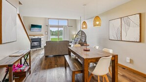 A spacious living room with a wooden dining table, a sofa, and a hanging chair. There is a fireplace, a large window with a view of greenery, and artwork on the walls. - Waterfront Boho Retreat (Wisconsin Dells)