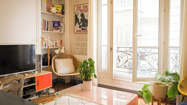A living room with a TV, bookshelf, and a large window with a balcony. - Chic Parisian Apartment in the Heart of the 17th (Paris)