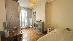 A bedroom with a wooden floor, a bed, a desk with a chair, a fireplace, and a window with a view of the outside. - Chic Parisian Apartment in the Heart of the 17th (Paris)
