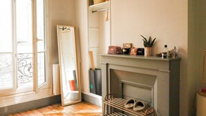 A room with a fireplace, a mirror leaning against the wall, a shoe rack, and a window with a view of the outside. - Chic Parisian Apartment in the Heart of the 17th (Paris)