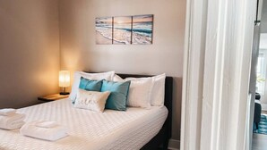 A neatly made bed with a white quilt, teal and white pillows, and a folded white towel. A wall-mounted painting of a beach scene is visible above the bed. - High Tide at CB - 75 steps to Beach Access (Carolina Beach)