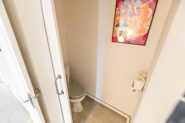 A small, narrow bathroom with a toilet, a wall-mounted toilet paper holder, and a poster on the wall.