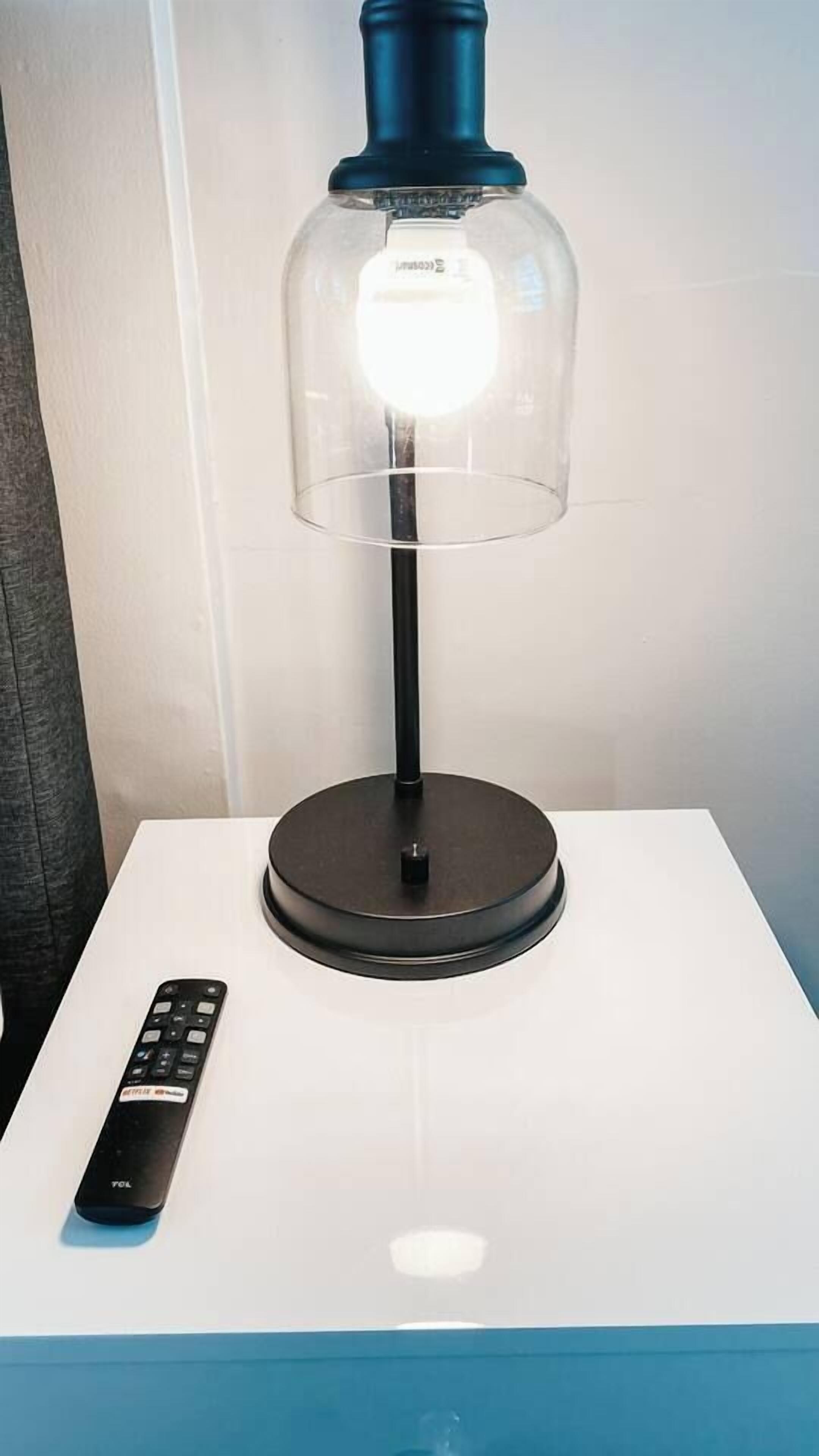 A table lamp with a clear glass shade and a black base, placed on a white table next to a remote control.