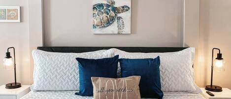 A neatly made bed with white and blue bedding, a turtle-themed artwork above it, and two bedside lamps.