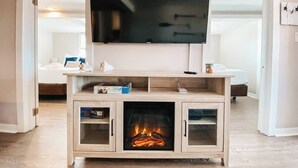 Een woonkamer met een aan een houten meubel bevestigde televisie, een open haard en een bed op de achtergrond.