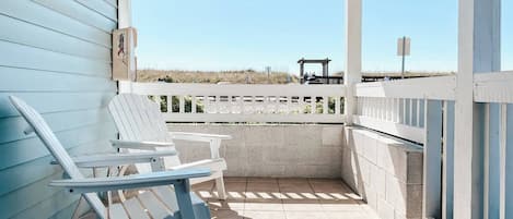 A covered outdoor seating area with two Adirondack chairs, a pergola with string lights, and a view of the beach.