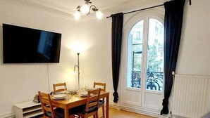A living room with a sofa, a dining table with chairs, a flat-screen TV mounted on the wall, and a window with curtains. - Haussmannian apartment at the gateway to Paris (Aubervilliers)