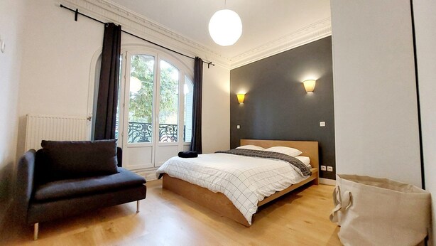 A bedroom with a bed, a chair, a window with curtains, and a wall-mounted light fixture. - Haussmannian apartment at the gateway to Paris (Aubervilliers)