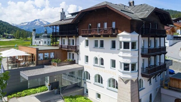 Ein mehrstöckiges Hotel mit einem markanten Balkon im oberen Stockwerk, freier Bergblick und einer gepflegten Außenbereich-Sitzgelegenheit.