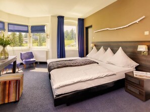 A hotel room with a large bed, a desk with a chair, a window with curtains, and a view of greenery outside. - Hotel Wetterstein (Seefeld in Tirol)