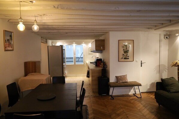 A cozy living room with a dining table, a sofa, and a bench. There is a kitchen area with cabinets and a refrigerator.