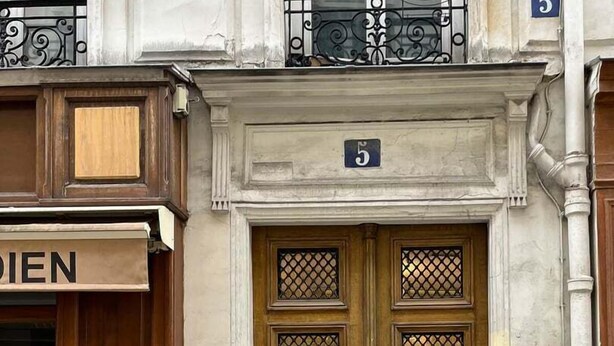 Un edificio con una puerta de madera doble, el número '5' encima y un cartel con la palabra 'DIEN' a la izquierda.