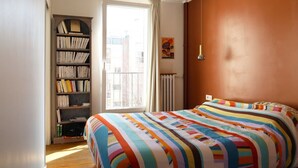 A bedroom with a large bed, a bookshelf, a window with curtains, and a radiator. - Garden View Apartment near Canal Saint-Martin (Paris)