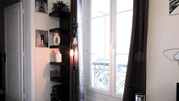 Un couloir avec une porte blanche, une fenêtre donnant sur un balcon et des étagères ornées d’objets décoratifs.