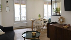 Een woonkamer met een donkere bank, een ronde tafel met een decoratieve schaal, een houten eettafel met stoelen, en een grote spiegel aan de muur.