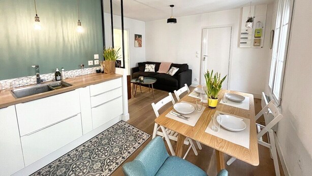 Een moderne keuken met witte kasten, een houten aanrecht en een spoelbak. Er is een eethoek met een houten tafel, stoelen en een potplant. Een zwarte bank en een klein rond tafeltje staan op de achtergrond.