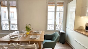 Een eetruimte met een houten tafel gedekt voor twee personen, een groene fauteuil en een prullenbak.