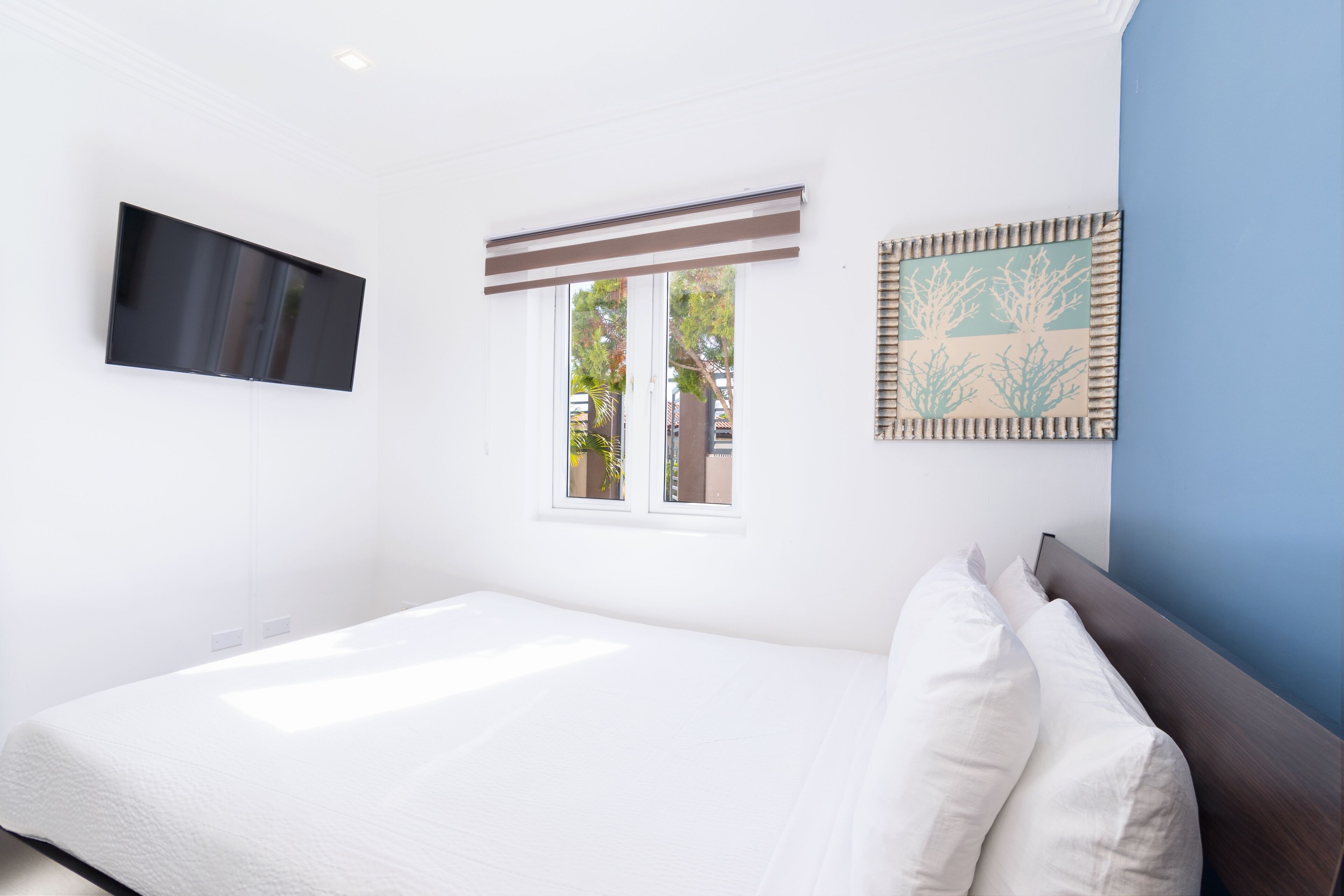 Une chambre à coucher avec un lit, un téléviseur fixé au mur, une fenêtre avec des stores et un tableau encadré accroché au mur.