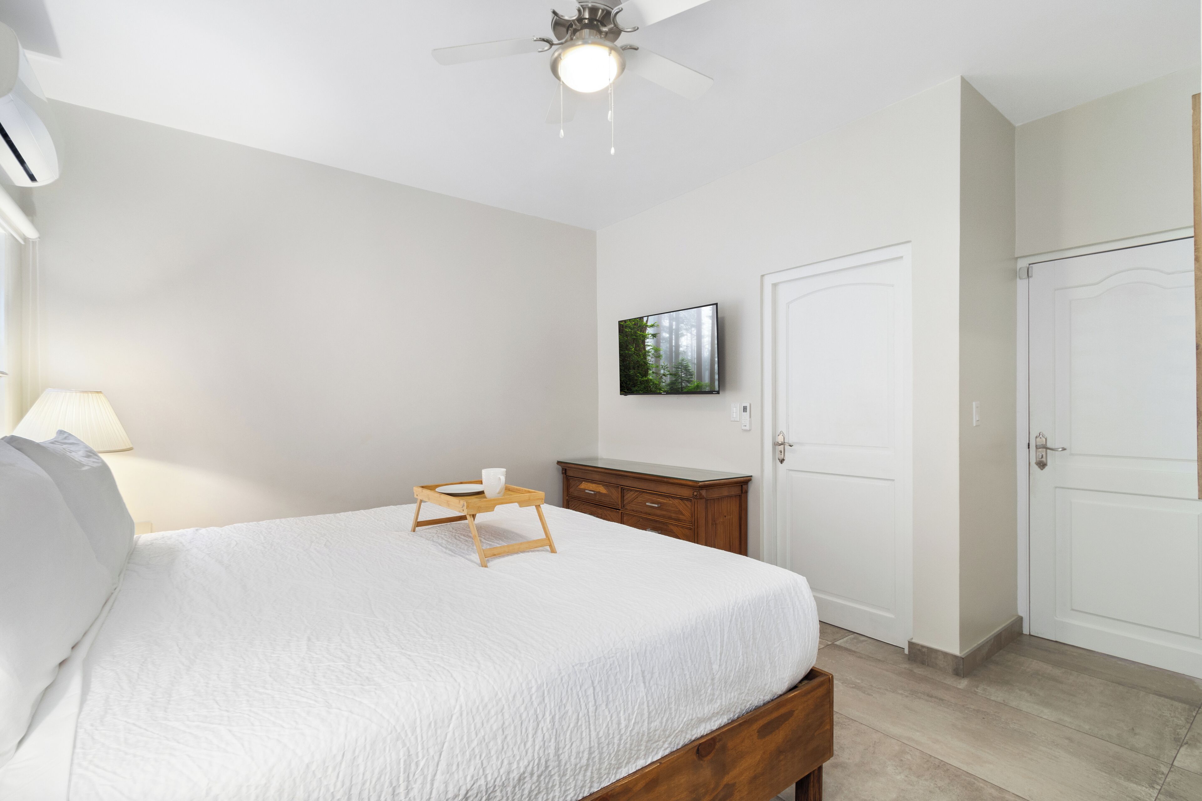 A bedroom with a bed, a bedside table with a lamp, a ceiling fan, a TV on the wall, and a wardrobe.