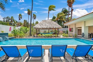 Un espace piscine avec des chaises longues bleues, une structure à toit de chaume et des palmiers.