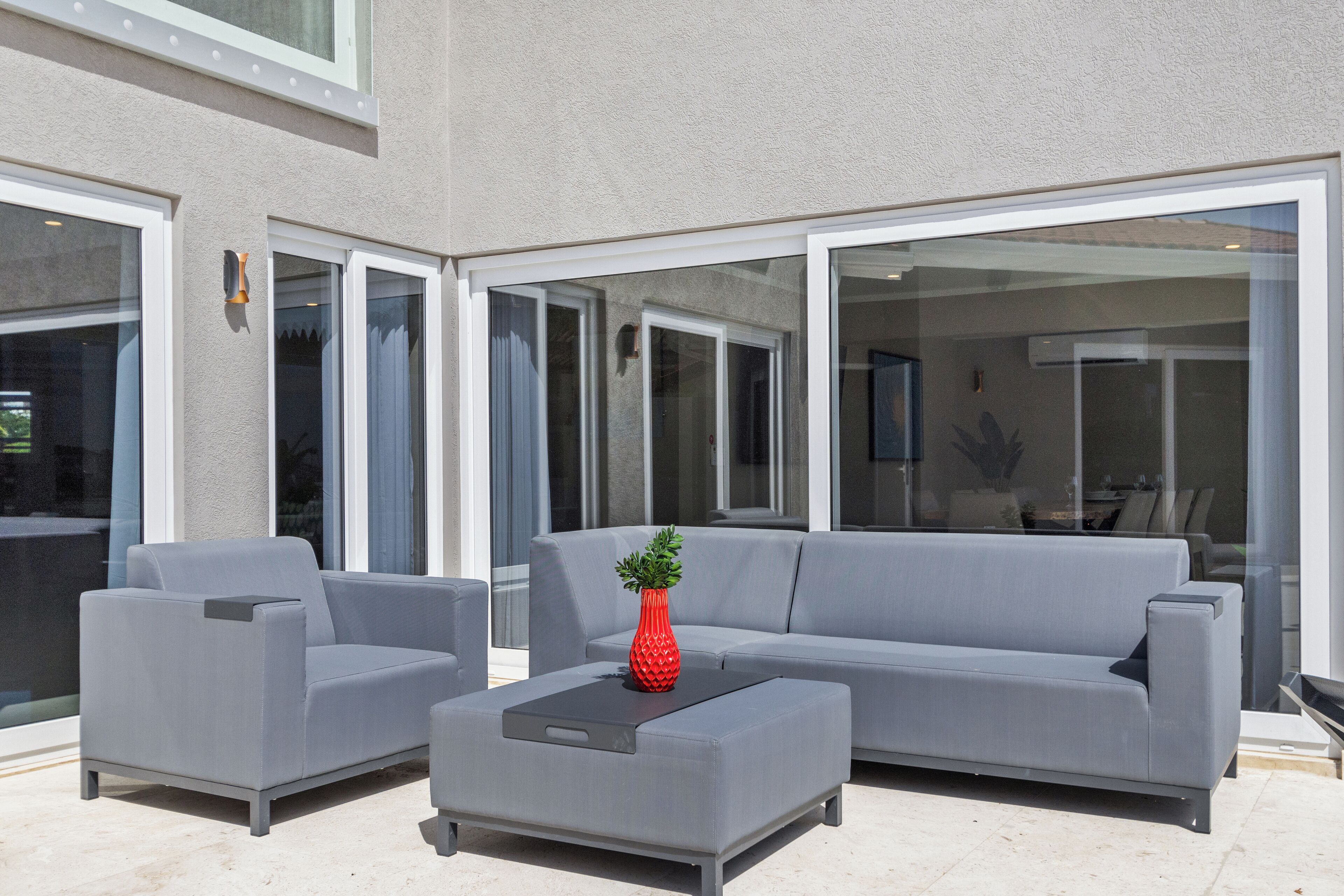 Un espace extérieur aménagé avec un canapé gris, des fauteuils, une petite table et un vase rouge.