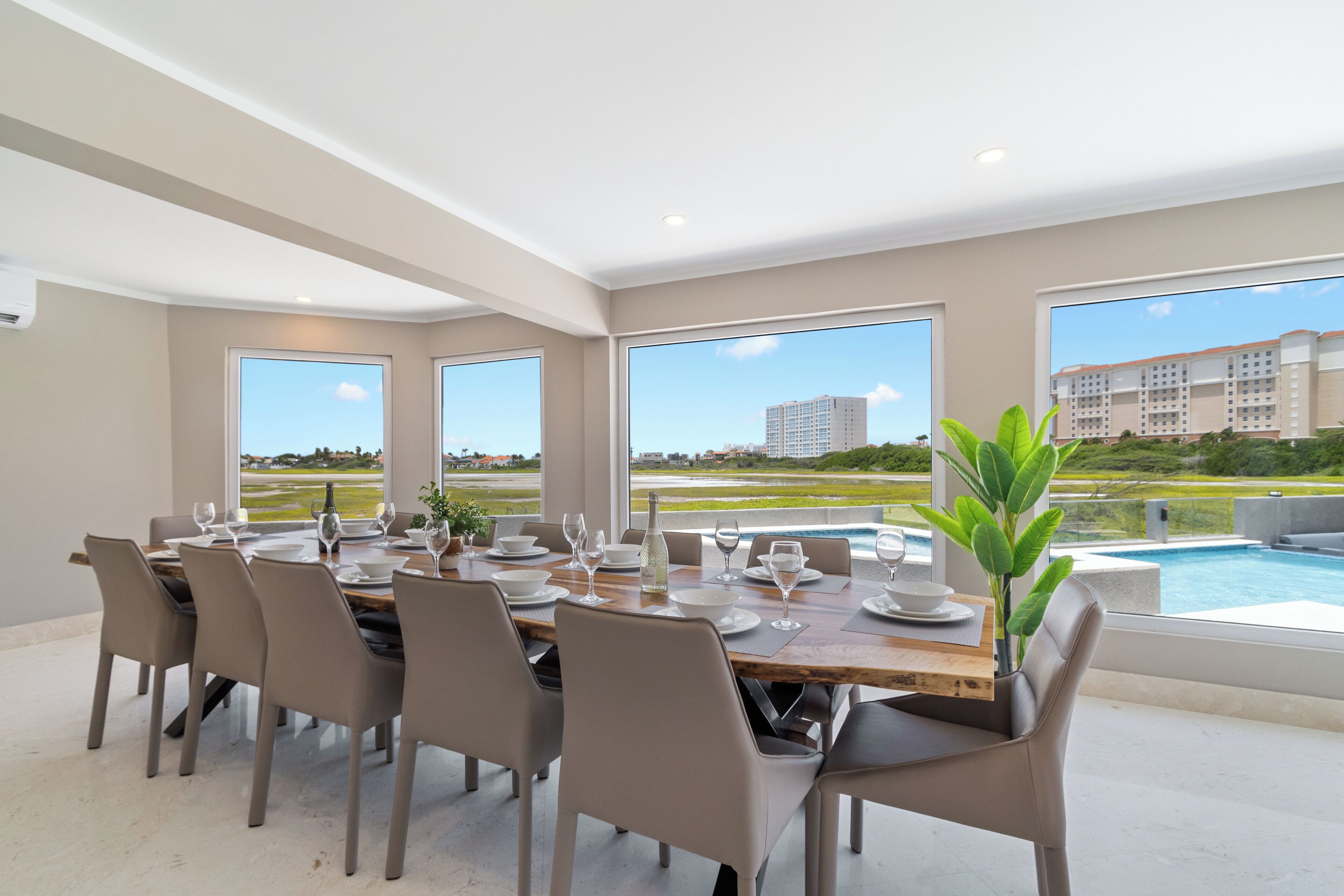 A dining area with a large table set for six, large windows, and a view of a swimming pool and buildings.