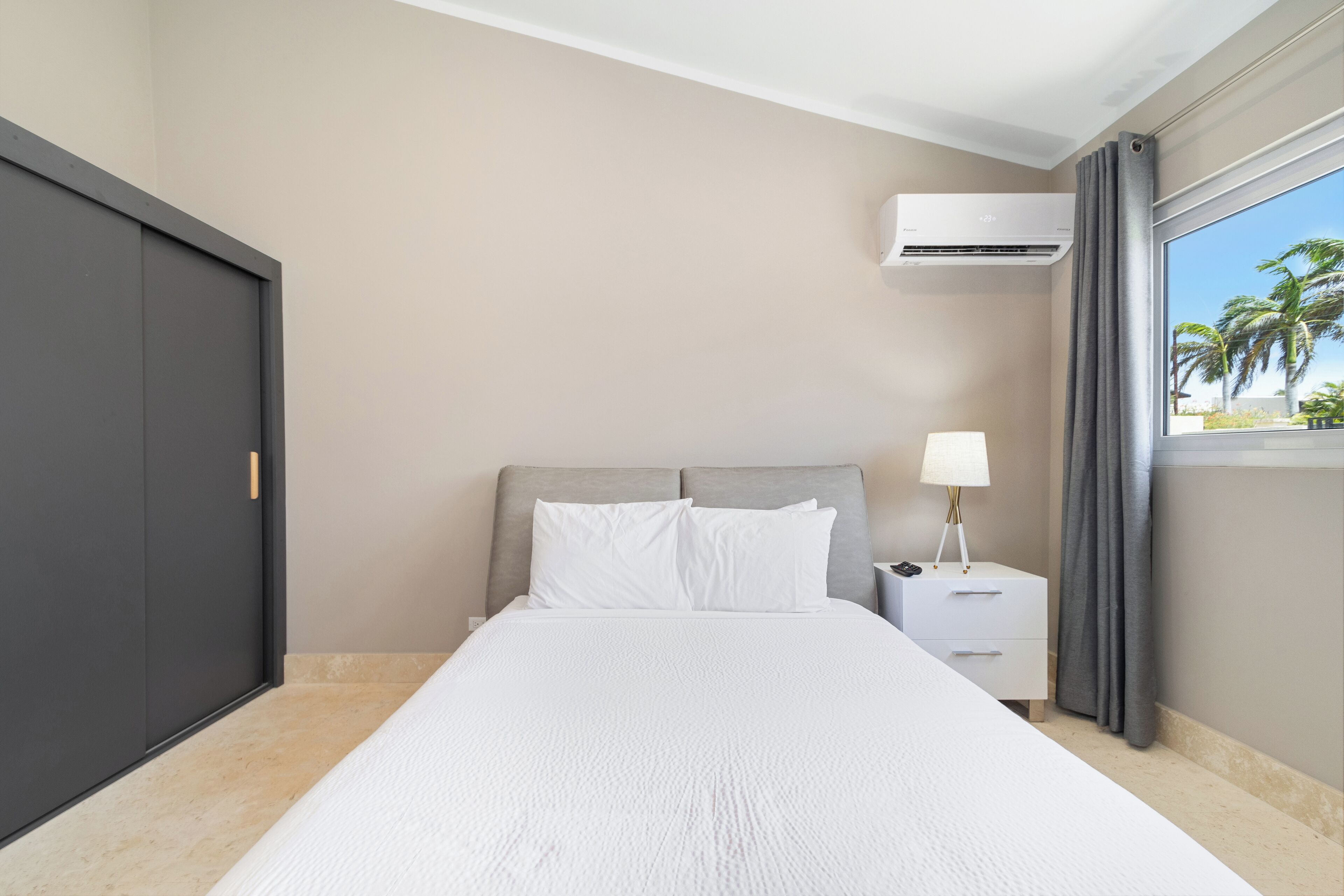A hotel room with a bed, a nightstand, a lamp, an air conditioning unit, and a window with curtains.