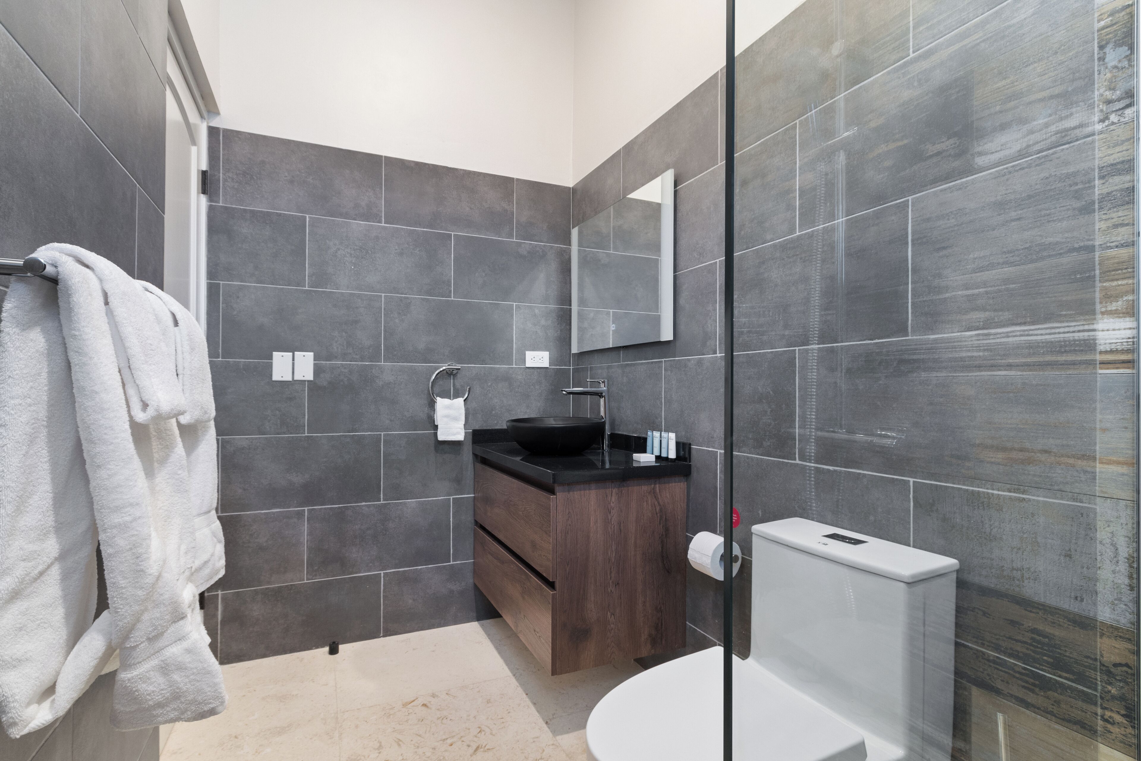 A modern bathroom with dark tiles, a wooden vanity, a black sink, and a glass shower enclosure.
