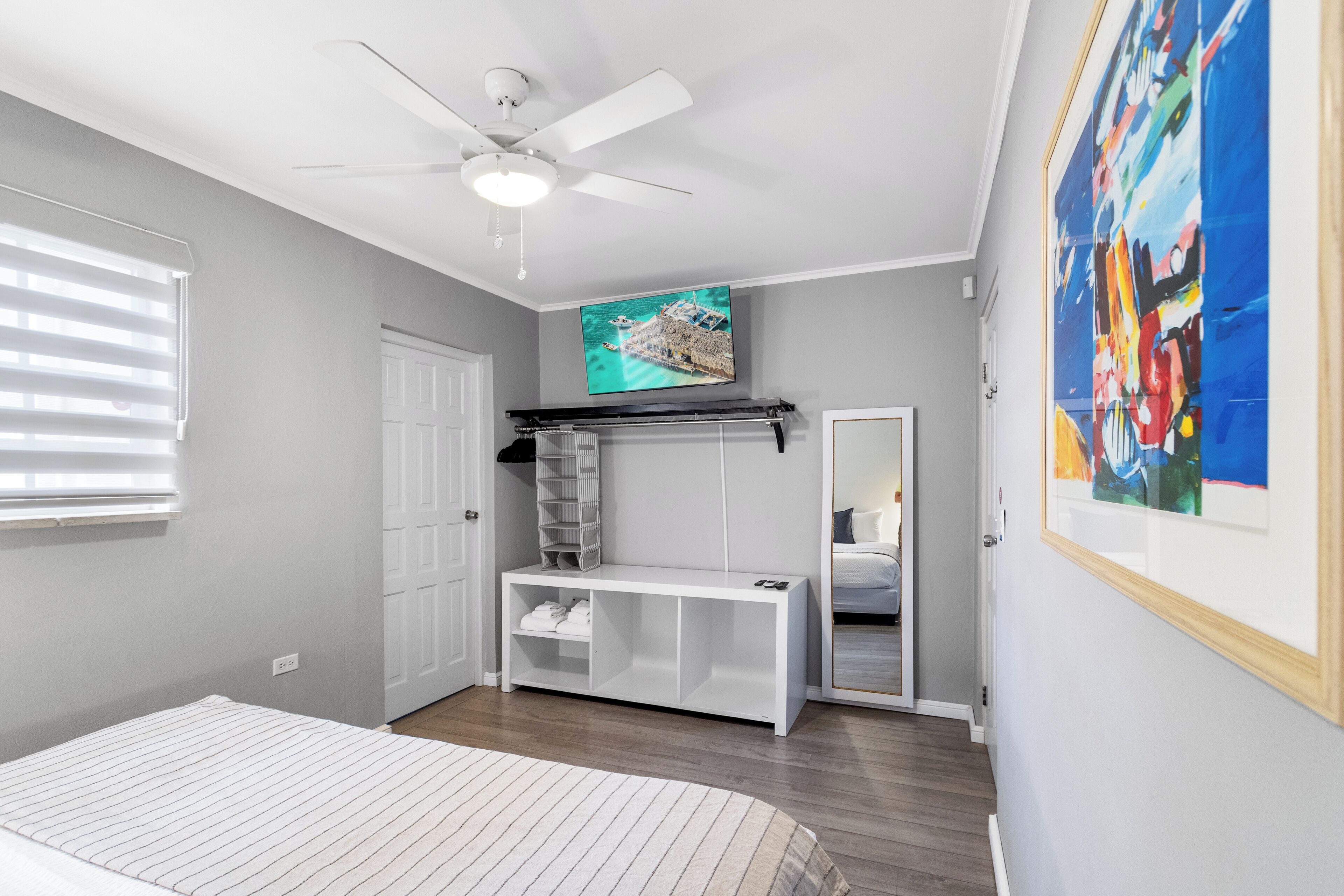 A bedroom with a bed, a ceiling fan, a TV, a bench, a door, and a painting.