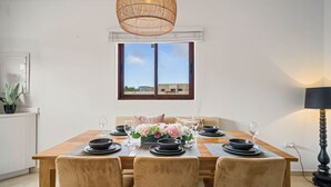A dining area with a wooden table set for four, a wicker pendant light, and a view of buildings through a window. - Felicidad 30 (Oranjestad)