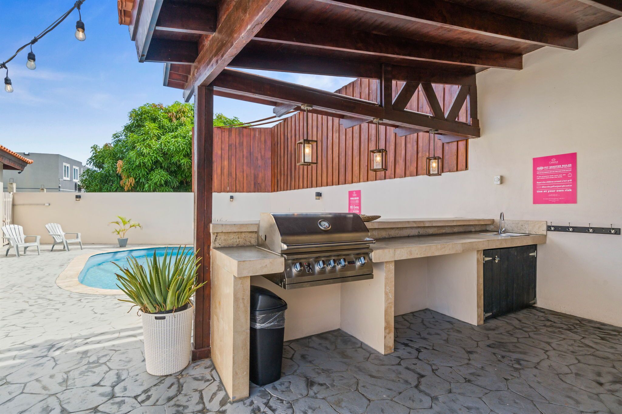 An outdoor kitchen with a grill, a sink, and a trash can. There is a pool in the background and a potted plant in the foreground.