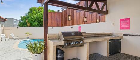 An outdoor kitchen with a grill, a sink, and a trash can. There is a pool in the background and a potted plant in the foreground.