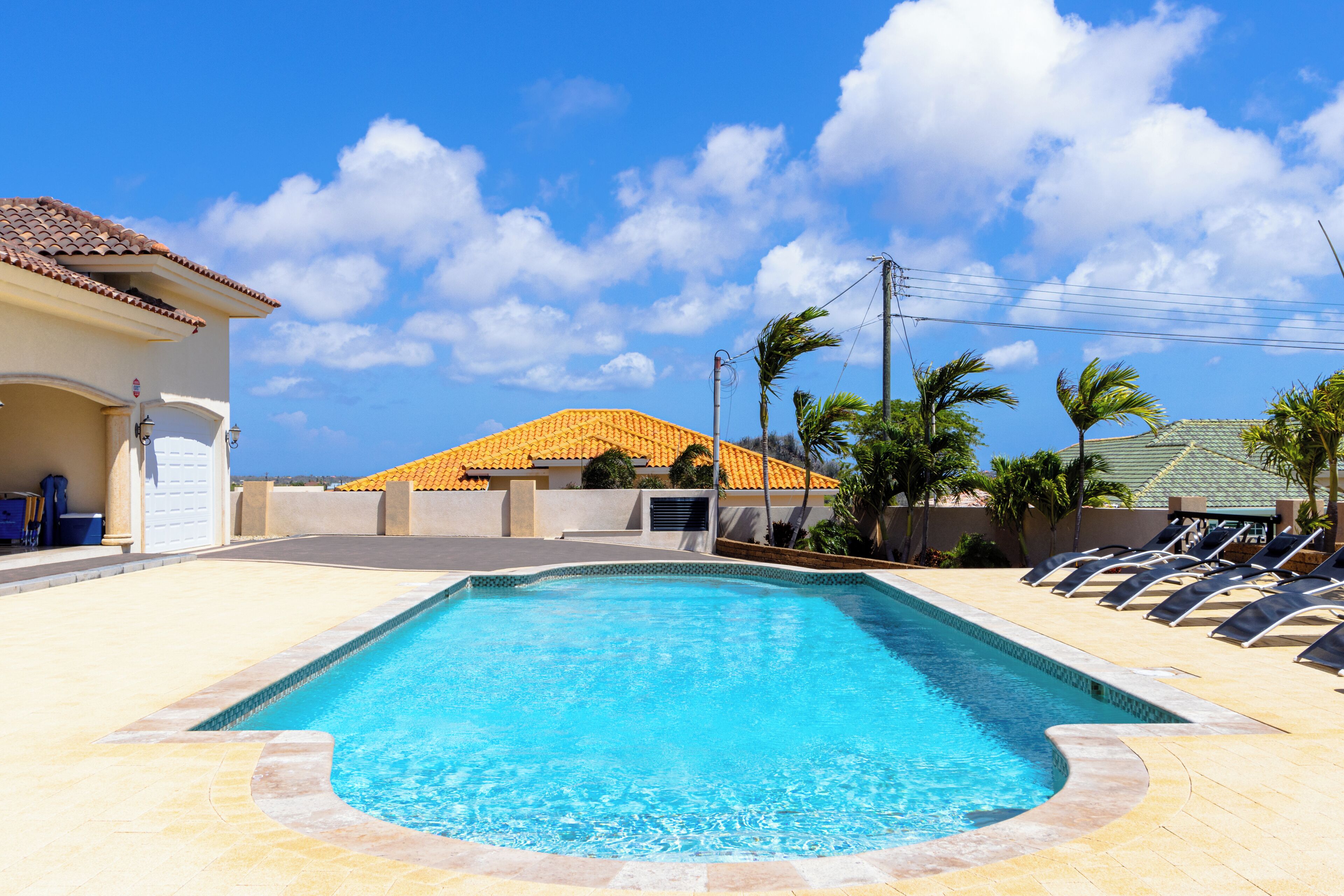 A swimming pool with a tiled deck, lounge chairs, and a building with a tiled roof in the background.