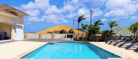 A swimming pool with a tiled deck, lounge chairs, and a building with a tiled roof in the background.