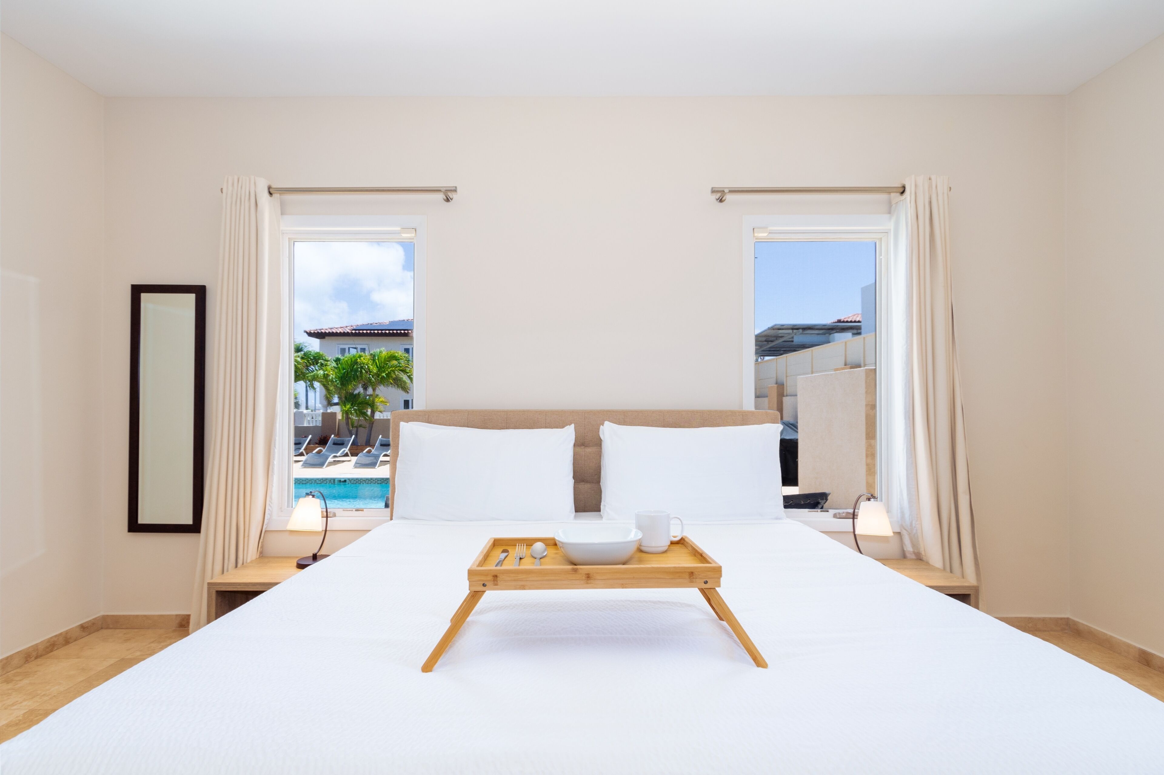 Une chambre d’hôtel avec un grand lit, un plateau avec des articles pour le petit-déjeuner, une vue sur une piscine et des bâtiments, et un tableau encadré au mur.