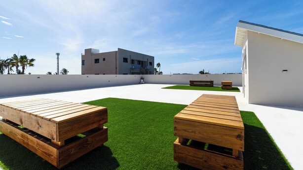 A modern outdoor seating area with wooden benches and a well-maintained lawn, adjacent to a white building with a flat roof. - Summerside 5 (Noord)