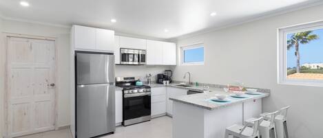 A modern kitchen with stainless steel appliances, a central island with bar stools, and a view of the beach through a window.